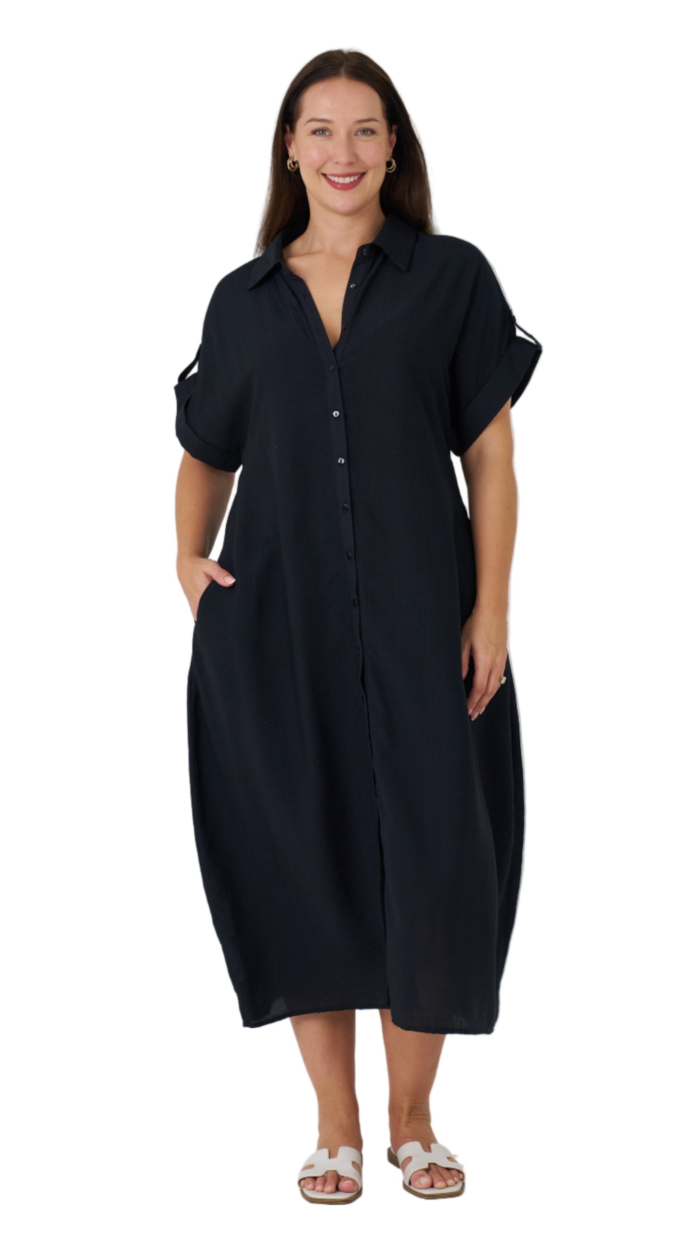 A person standing in a studio setting, wearing a black midi length dress with a relaxed silhouette, button-down front, and roll-up sleeves.