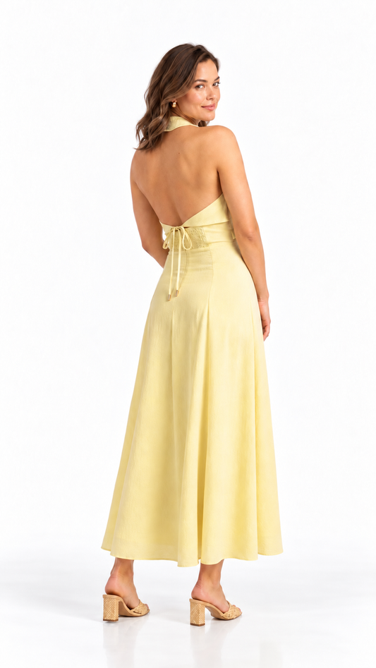 A woman modeling a pale yellow halter-neck maxi dress from the back, featuring a tie waist and open back, paired with beige block-heel sandals on a white studio background.