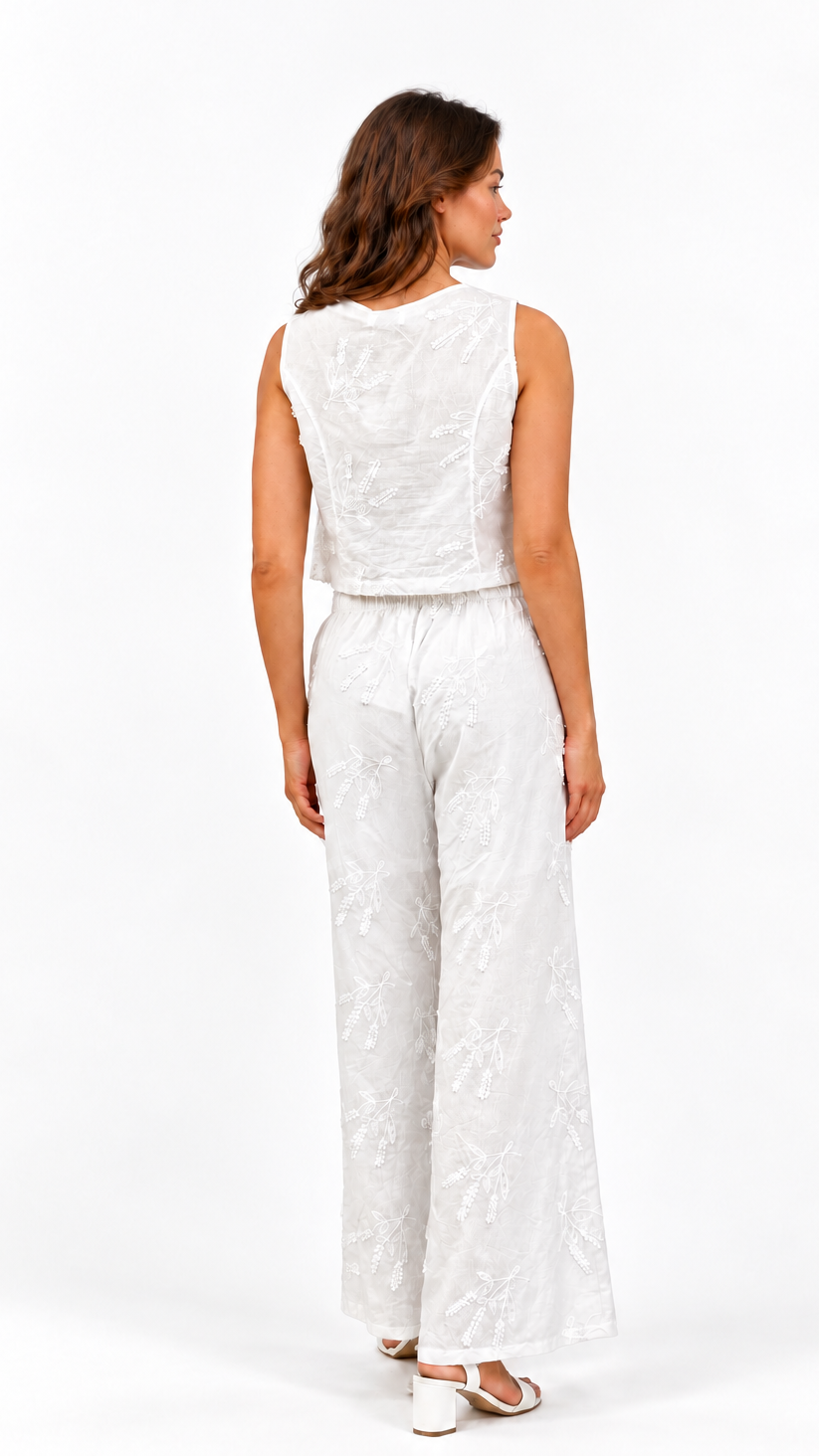 A woman viewed from the back wearing a sleeveless white textured jumpsuit with wide-leg pants and white heeled sandals, photographed against a clean white studio background.