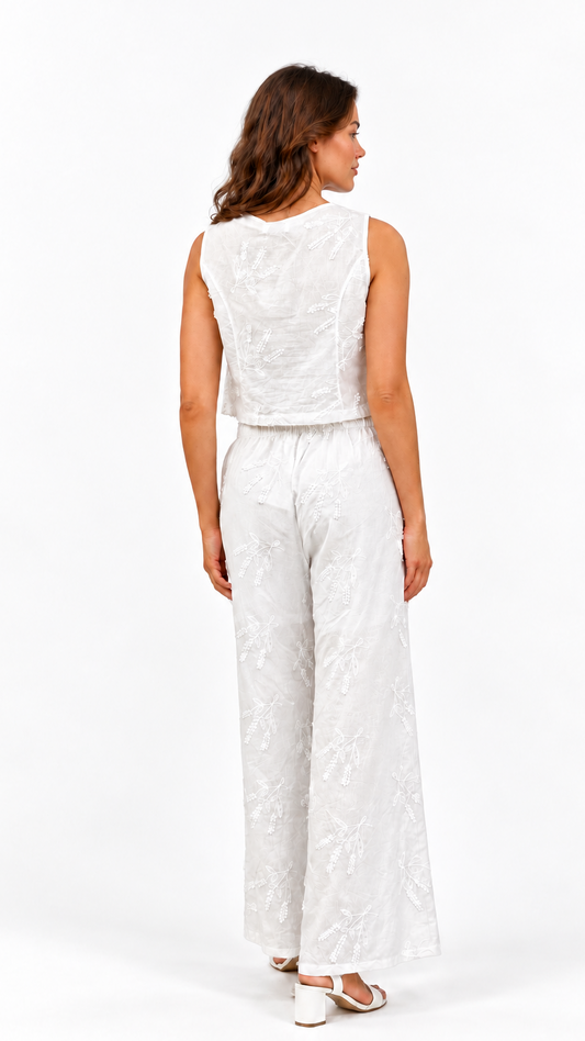 A woman viewed from the back wearing a sleeveless white textured jumpsuit with wide-leg pants and white heeled sandals, photographed against a clean white studio background.