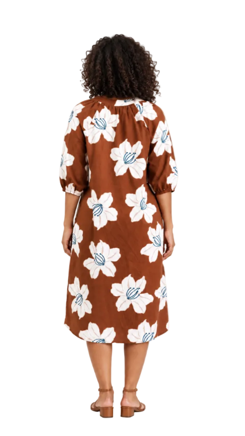 A woman viewed from behind wearing a brown midi dress with a large white floral print and light blue center details, paired with brown heeled sandals, against a plain white studio background.