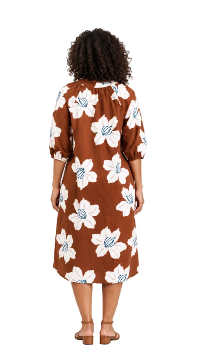A woman viewed from behind wearing a brown midi dress with a large white floral print and light blue center details, paired with brown heeled sandals, against a plain white studio background.