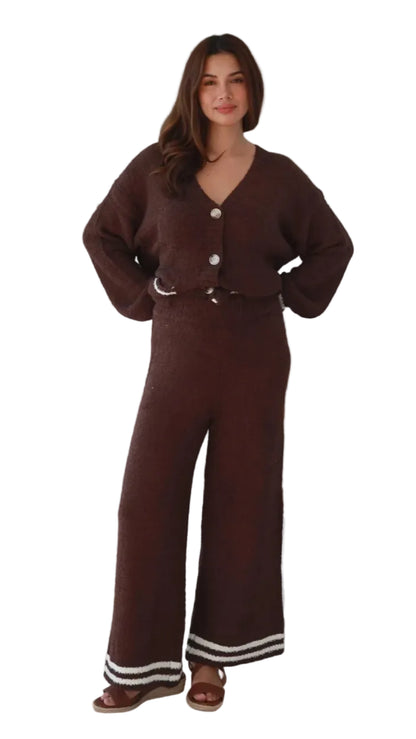 A woman modeling a dark brown knit matching set consisting of a button-front cardigan and wide-leg pants with white striped trim at the hems, shown against a plain white background.