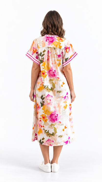 A woman viewed from behind wearing a short-sleeve midi dress with a colorful floral print on a cream background, featuring pink and white striped trim on the collar and sleeve cuffs, paired with white sneakers against a white studio background.