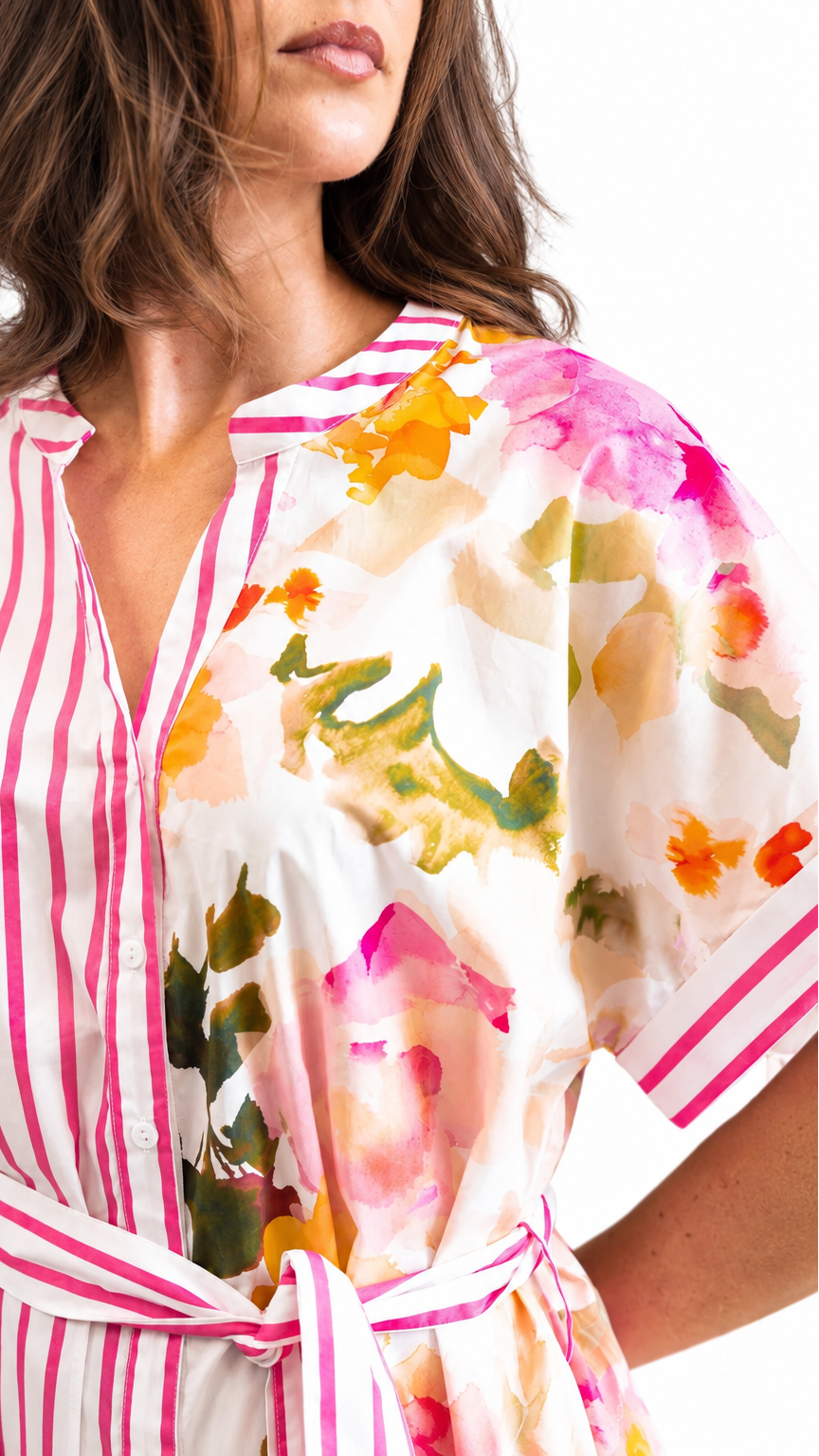 A close-up of a woman wearing a short-sleeve shirt dress with a split design: one side has pink and white vertical stripes, and the other side has a white base with a colorful watercolor floral print in pink, orange, yellow, and green. The dress has a V-neck and a matching tie belt at the waist.