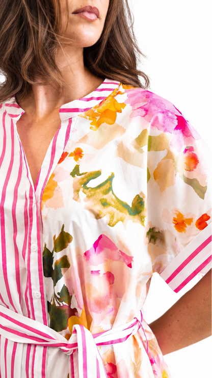 A close-up of a woman wearing a short-sleeve shirt dress with a split design: one side has pink and white vertical stripes, and the other side has a white base with a colorful watercolor floral print in pink, orange, yellow, and green. The dress has a V-neck and a matching tie belt at the waist.