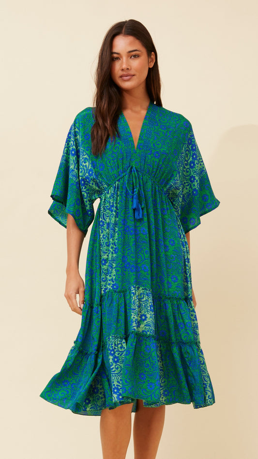 Woman wearing a vibrant blue and green floral Esmerelda Midi Dress with tassel tie and tiered hem – front view.