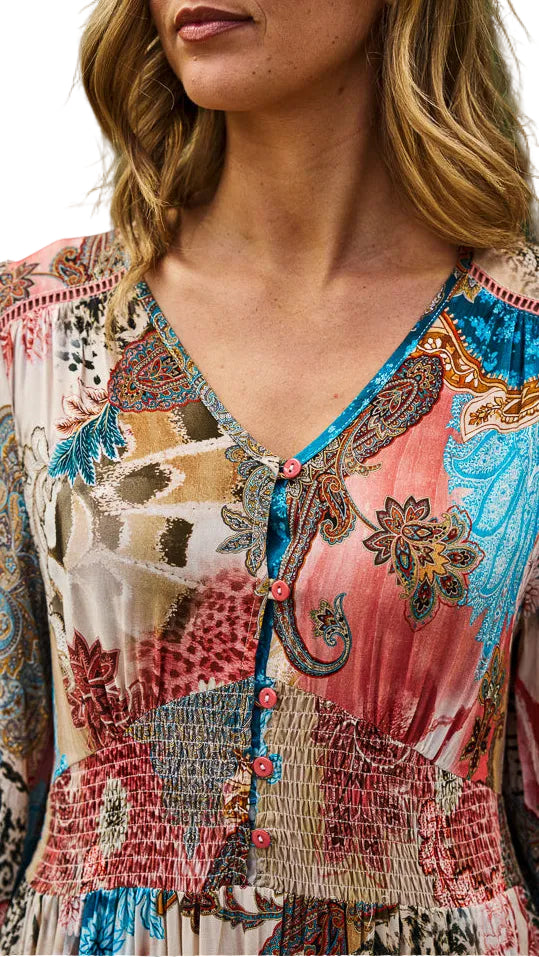 A close-up of a woman wearing a bohemian-style long-sleeve dress with a multicolor paisley and floral print in pink, blue, beige, and brown tones, featuring a smocked waist and button-front neckline.