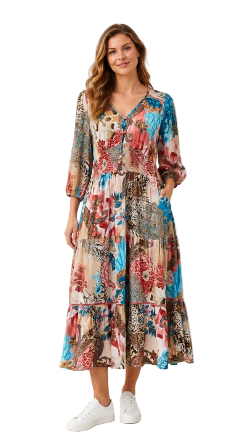 A woman modeling a long-sleeved, tiered midi dress with a vibrant multicolor floral and paisley print, paired with white sneakers, against a plain white background.