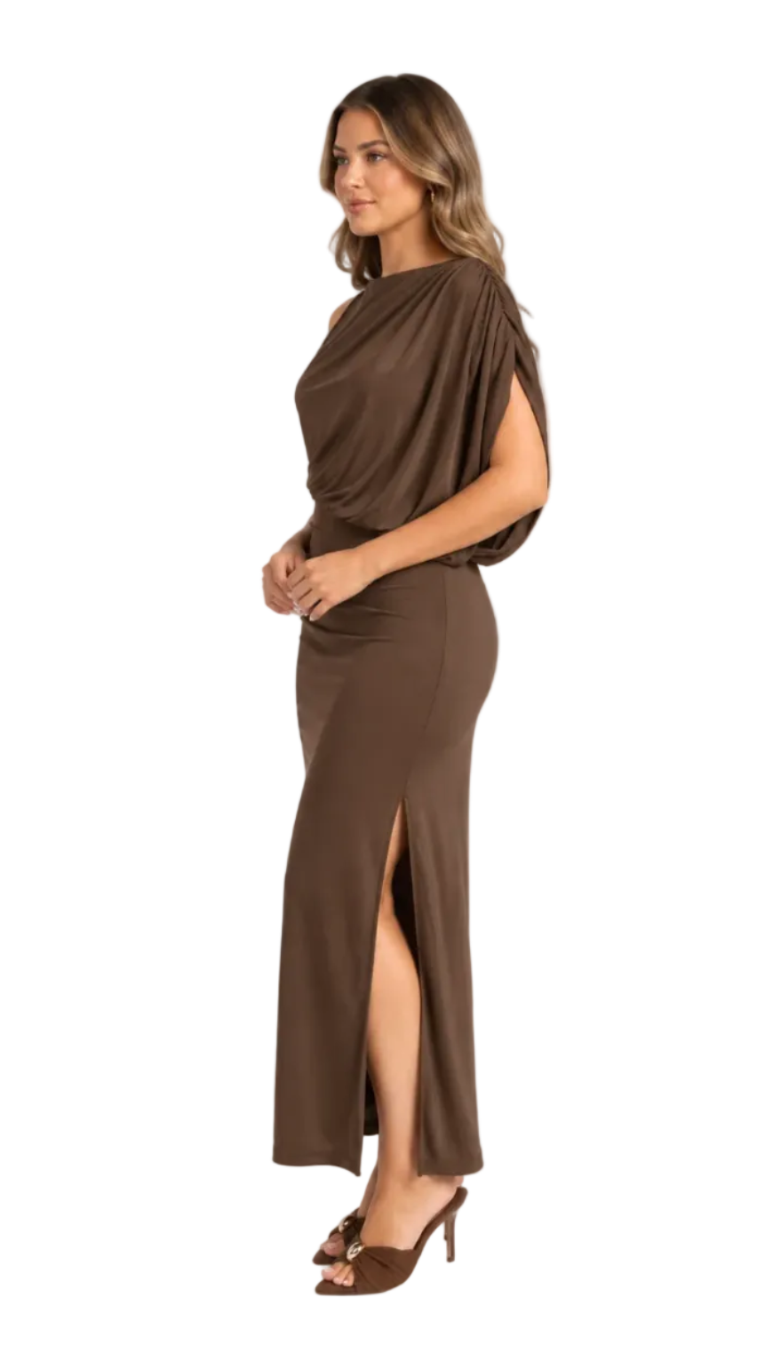 A woman modeling a floor-length brown one-shoulder draped maxi dress with a thigh-high slit, paired with brown heeled sandals, against a light gray studio background.