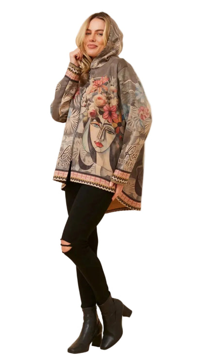 A woman modeling a hooded poncho-style jacket with a detailed graphic print of a woman's face and floral elements, styled with black ripped leggings and black ankle boots against a plain white background.