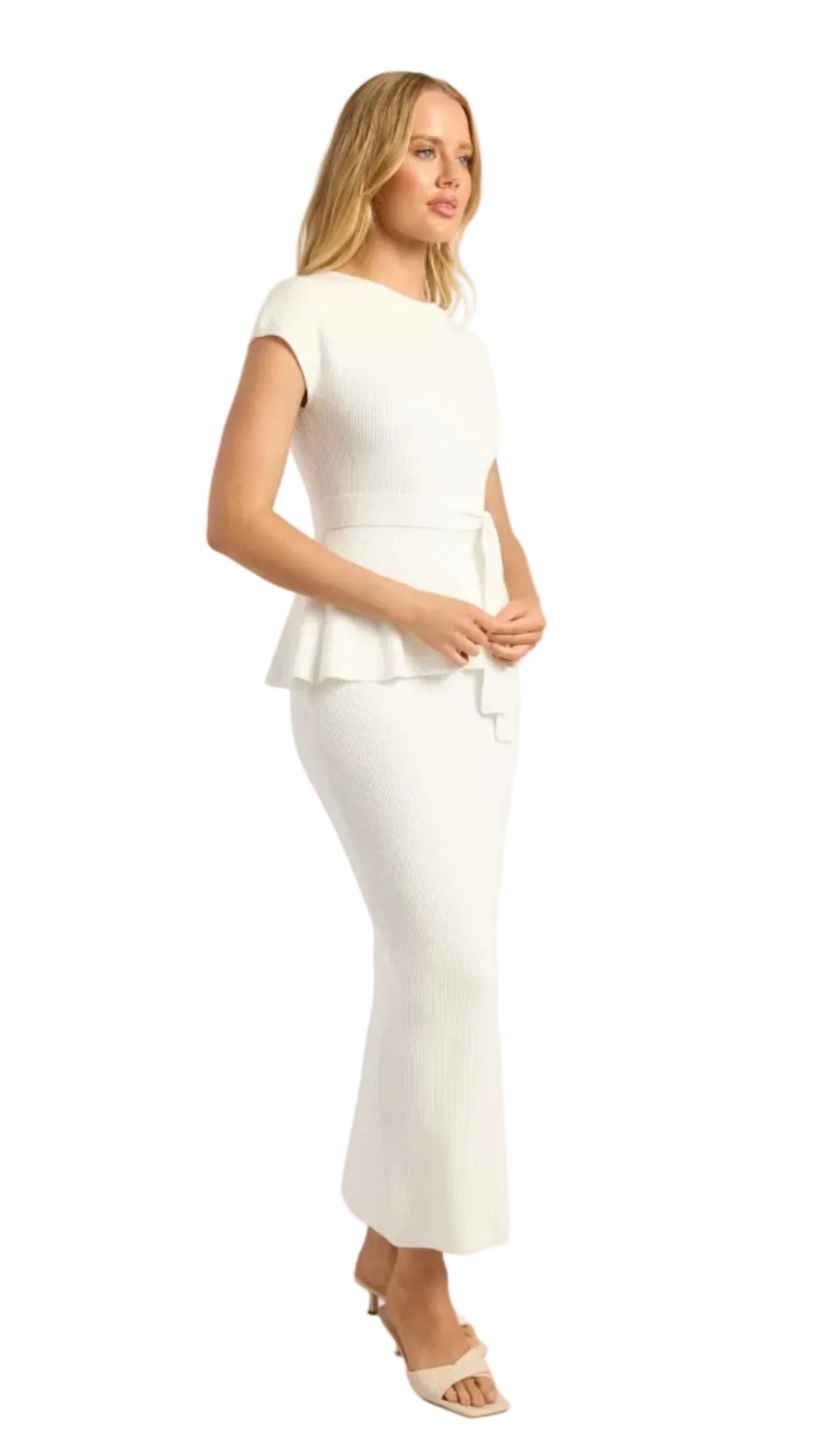 A woman modeling a white sleeveless peplum midi dress with a matching tie belt, paired with beige heeled sandals, against a light gray studio background.