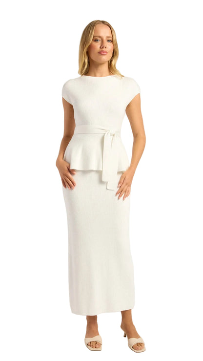 A woman modeling a white two-piece knit set consisting of a short-sleeve peplum top with a tie waist and a matching midi skirt, paired with beige heeled sandals against a light gray studio background.