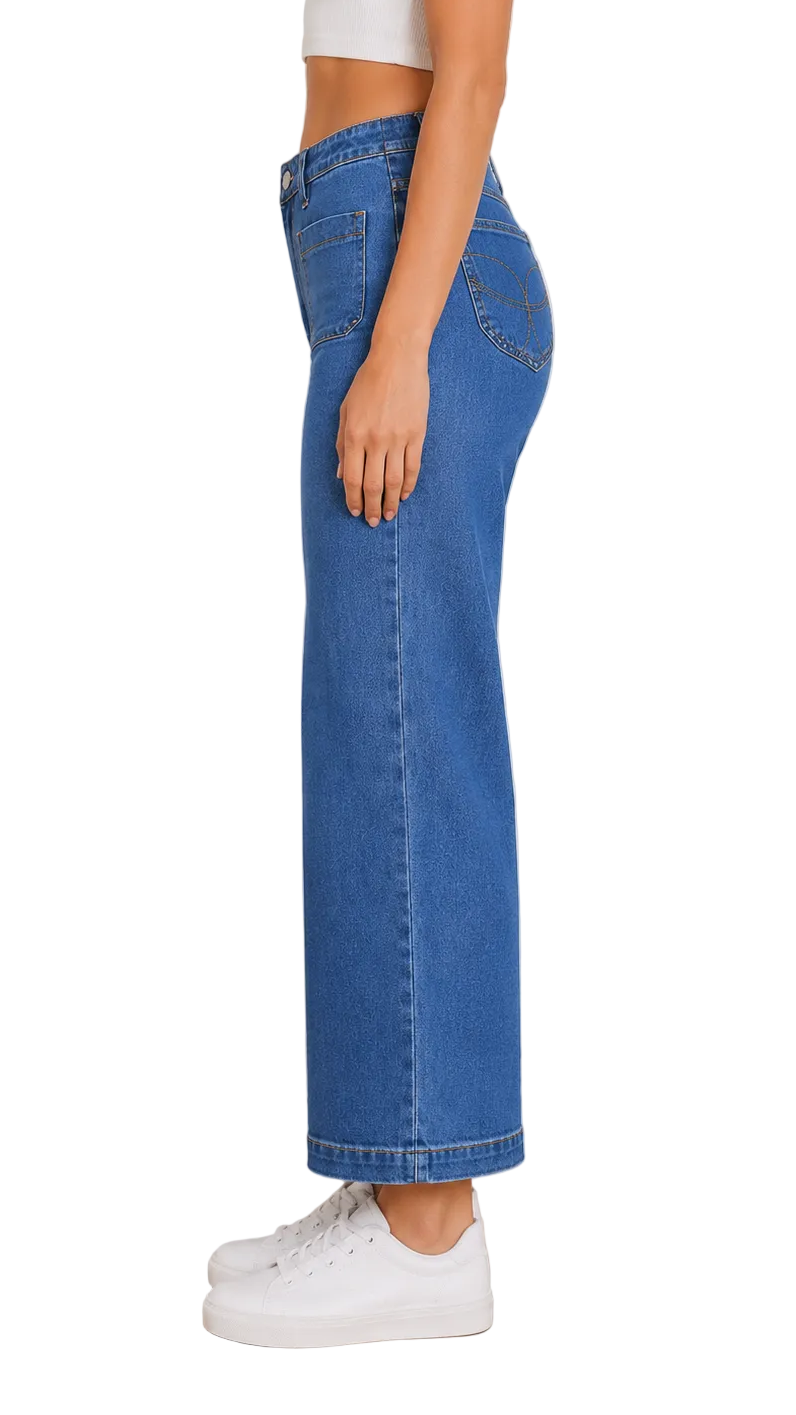 A side-profile view of a model wearing high-waisted, wide-leg blue denim jeans with a cropped white top and white sneakers, set against a neutral indoor background.