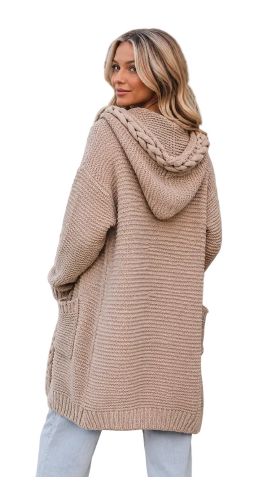 A woman wearing a chunky knit hooded cardigan in a warm taupe-beige color, standing outdoors in front of green palm leaves and a white wall.