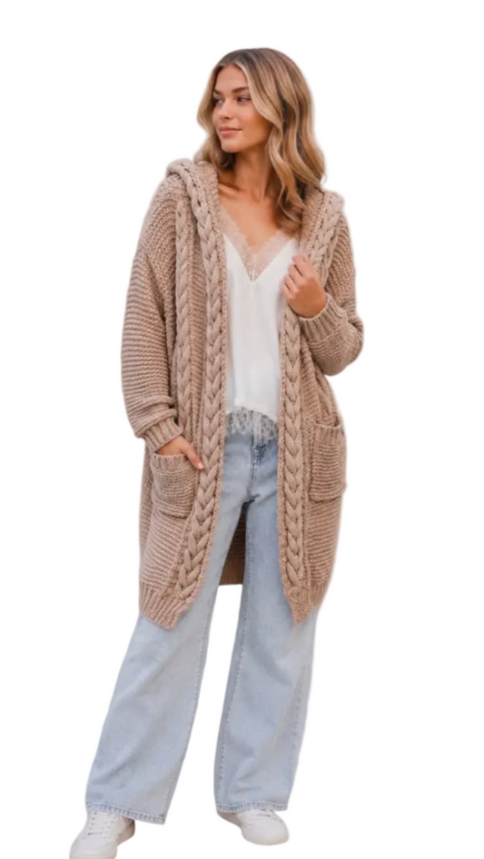 A woman wearing a chunky, long-sleeve beige cable-knit cardigan with a hood and front pockets, styled over a white top and light-wash jeans, 