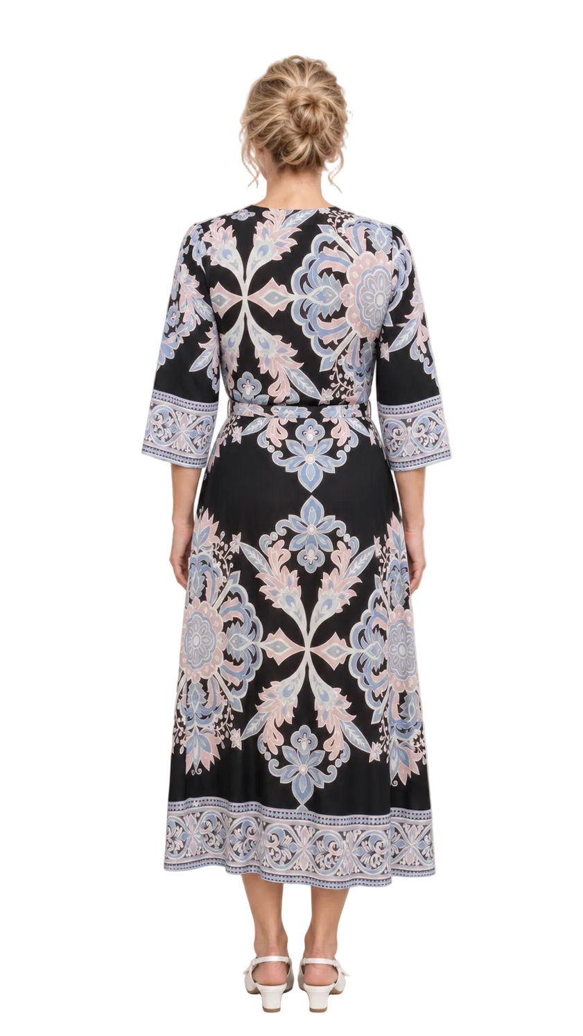 A woman viewed from the back wearing a black midi dress with ornate light blue and pale pink floral embroidery and a matching embroidered hem, paired with white heeled sandals against a neutral beige studio backdrop.
