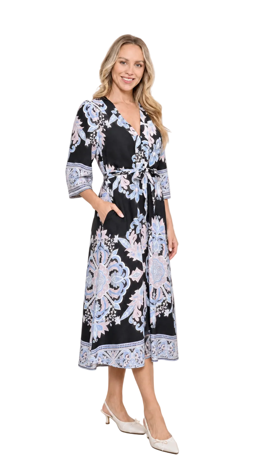 A woman modeling a black midi wrap dress with a light blue and white floral pattern, featuring elbow-length sleeves, a V-neckline, and a self-tie belt, set against a neutral beige studio backdrop.